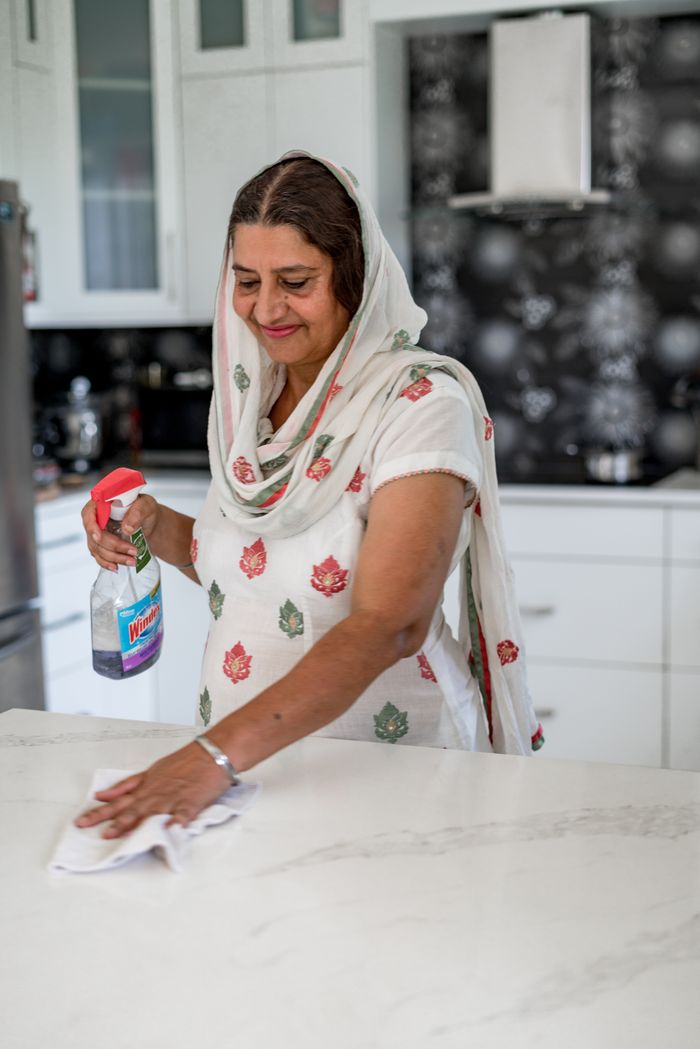 Senior woman wearing headscarf wipes kitchen counter with cleaning spray. Indoor setting.